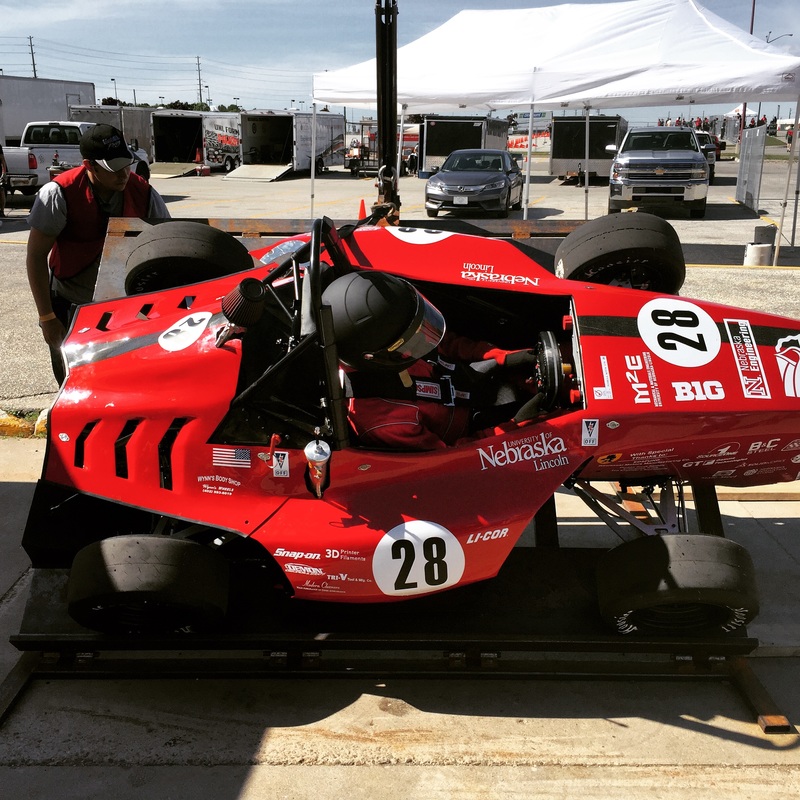 Red race car with #28 being inspected on a platform in a sunny outdoor setting.