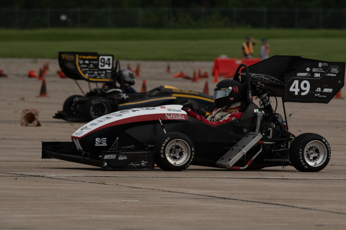 Race cars with labeled numbers competing on a wide asphalt track surrounded by orange cones.