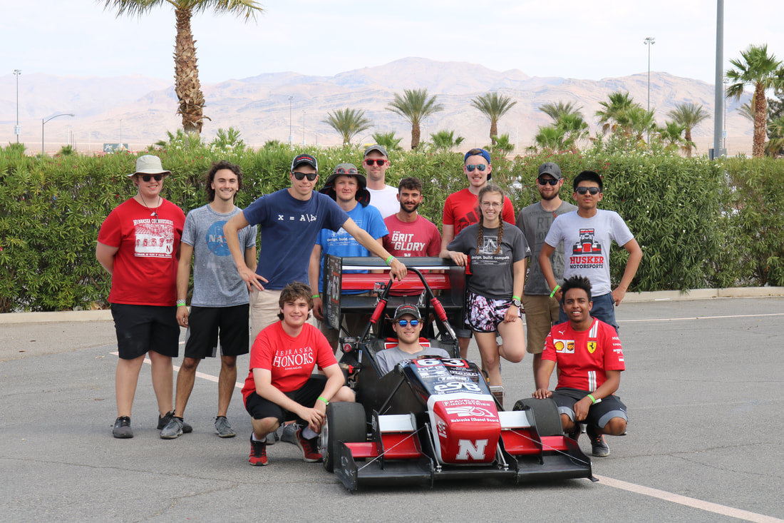 Group posing outdoors with a red race car in front, palm trees and mountains in the background.