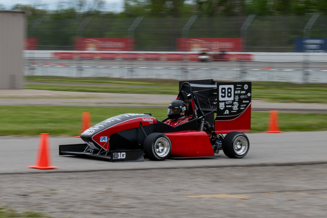 Red and black race car speeding on a track with orange cones nearby.