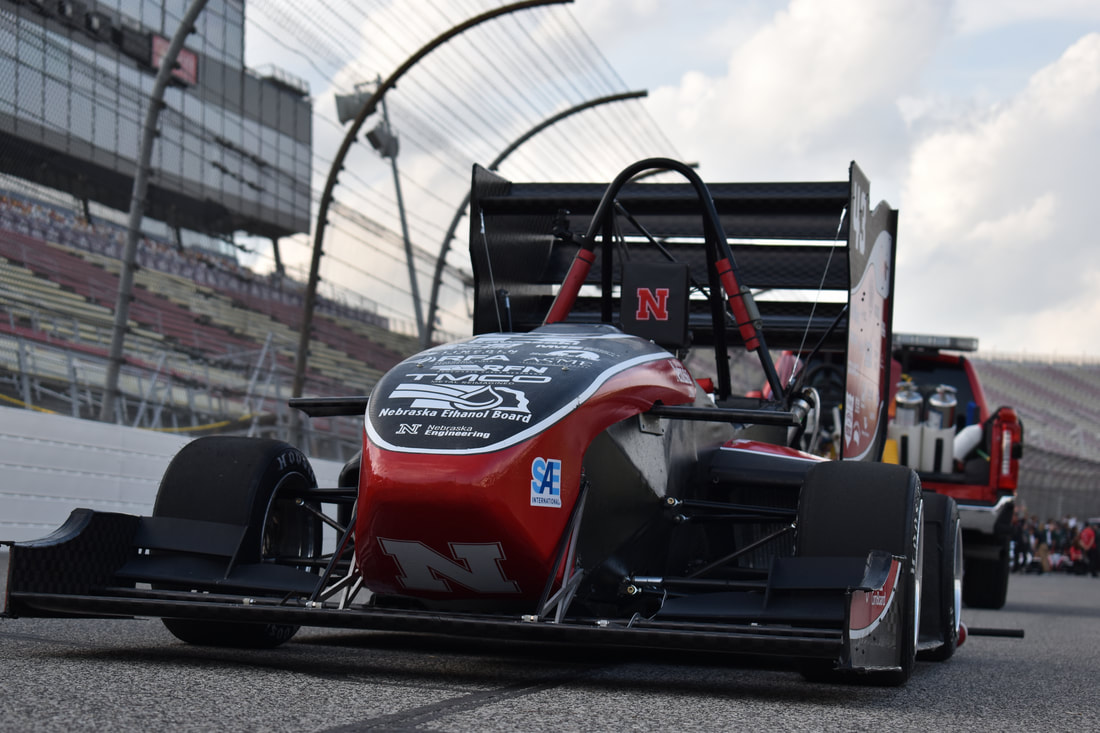 Red and black race car on a track, surrounded by stadium seating and a truck in the background.
